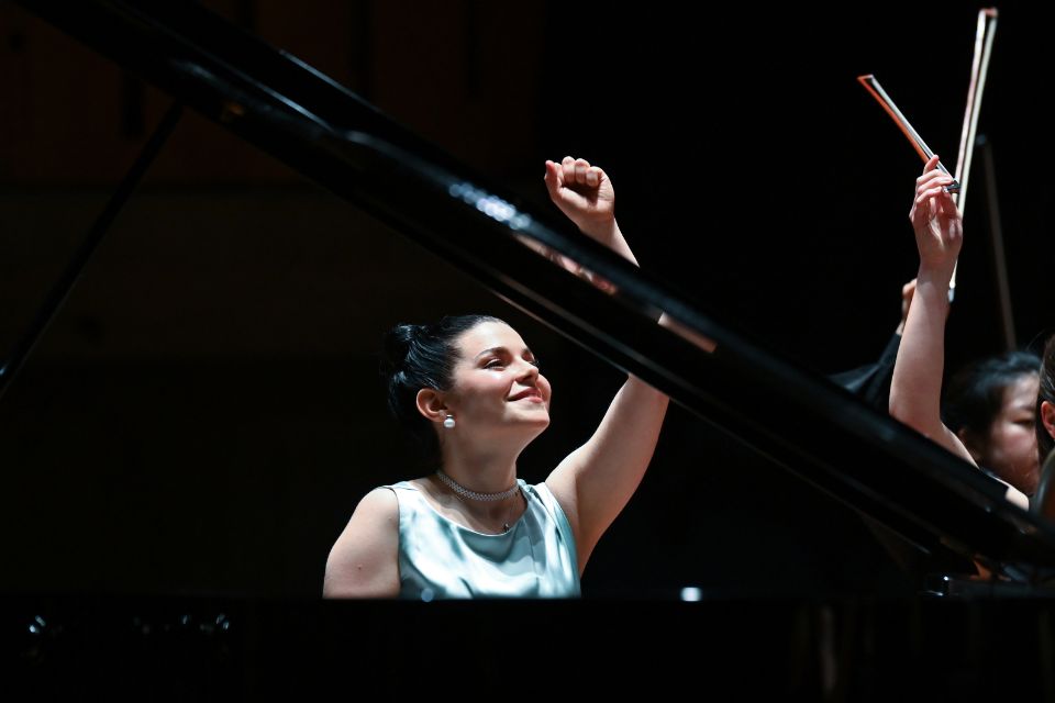 A woman wearing a silver dress and black hair tied up sat behind a piano with one arm raised in the air, smiling.